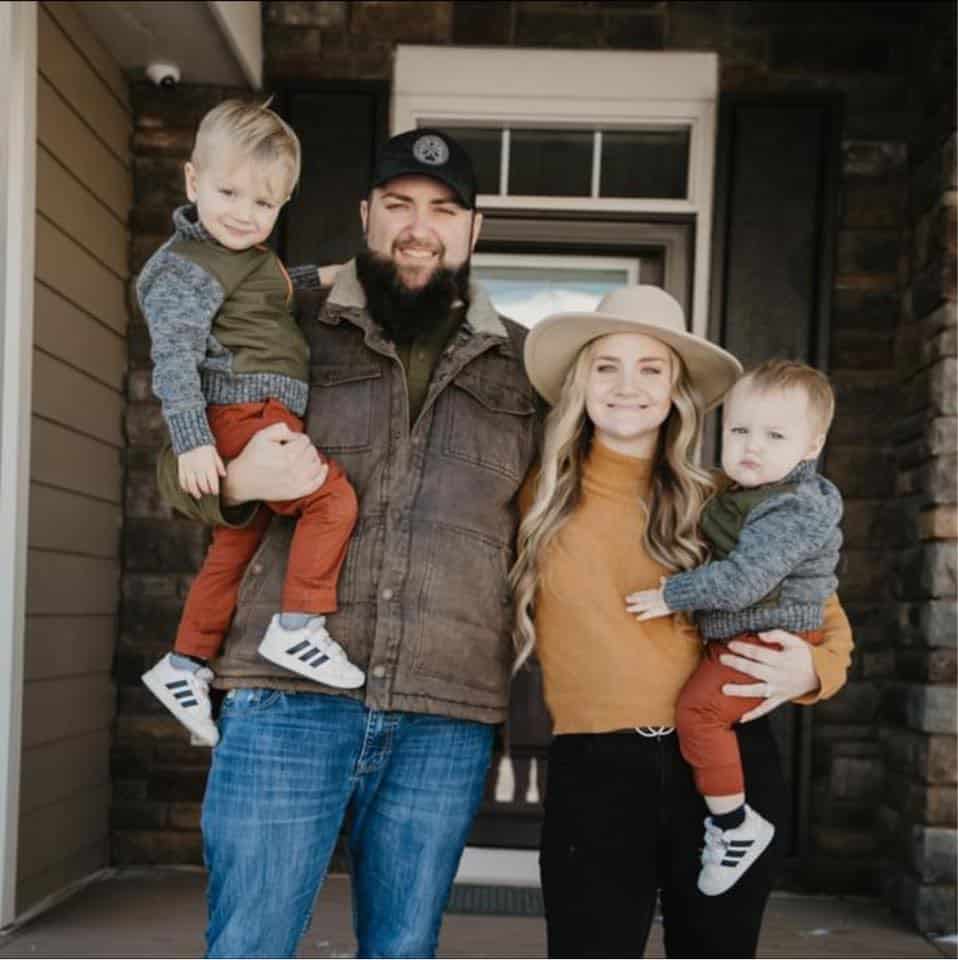 A family portrait in front of a house. Sam Romain holding a young child on his hip, stands next to a woman wearing a hat, who is holding a toddler. They are both smiling at the camera.