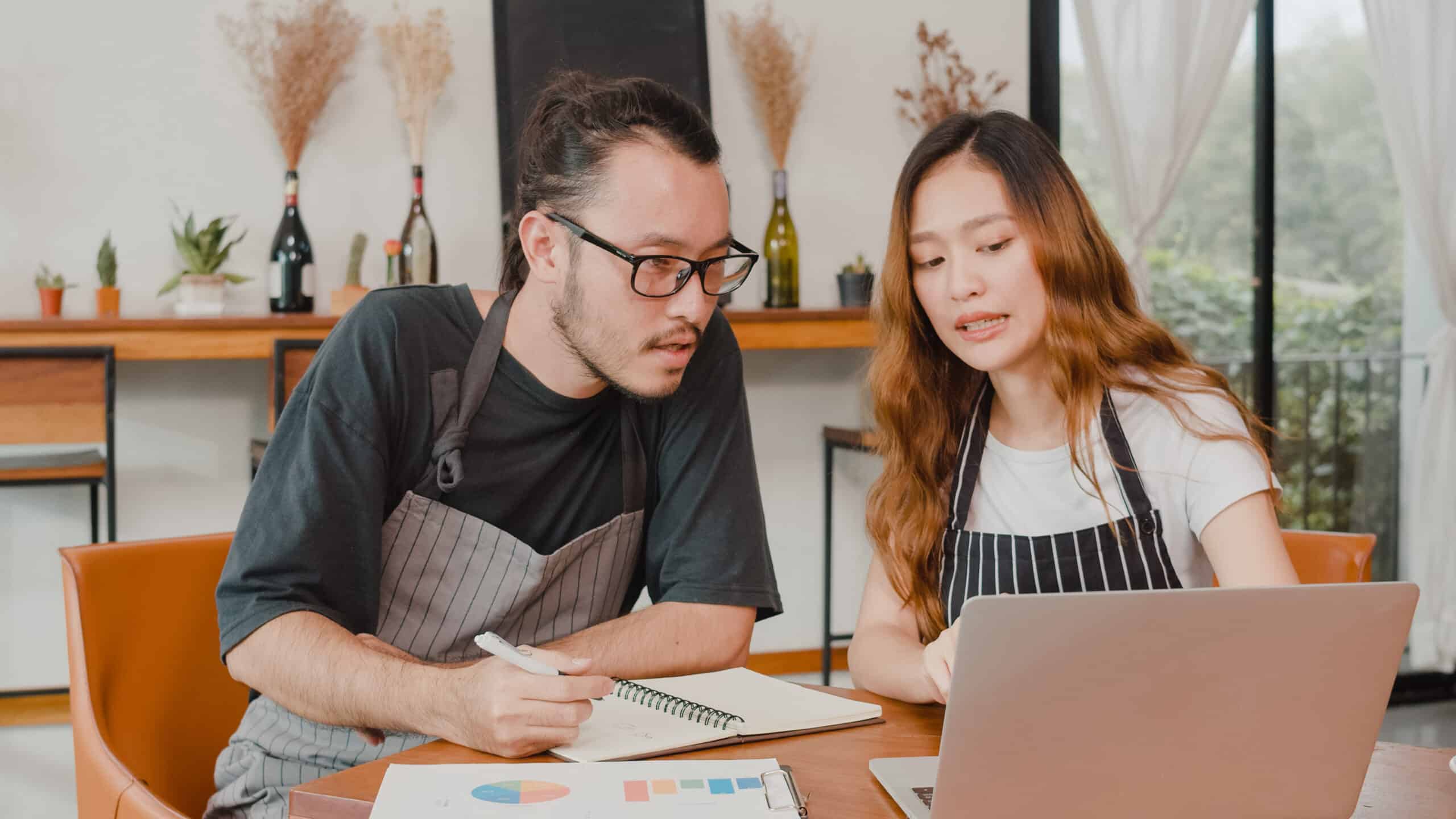 Stressed Asian small business owners couple using laptop discussing project and finance with documents of shop for the month at cafe.