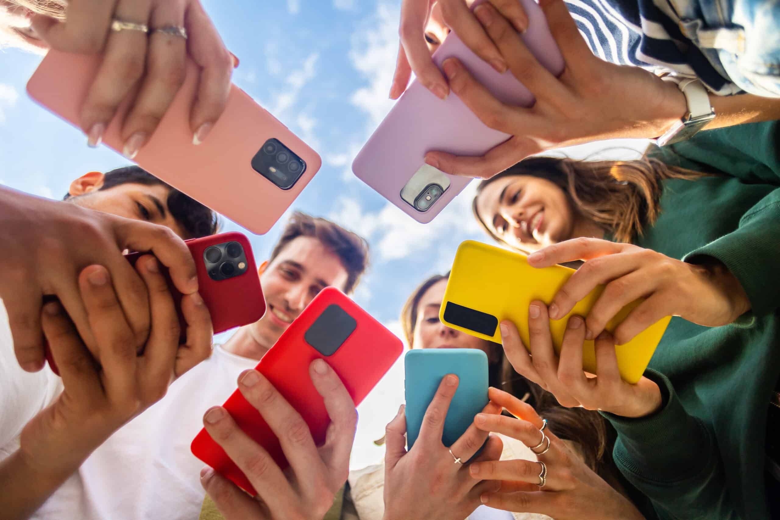 Young group of people using mobile phone device standing together in circle outdoors. Millennial friends addicted to social media app, betting or playing video game on platform online.