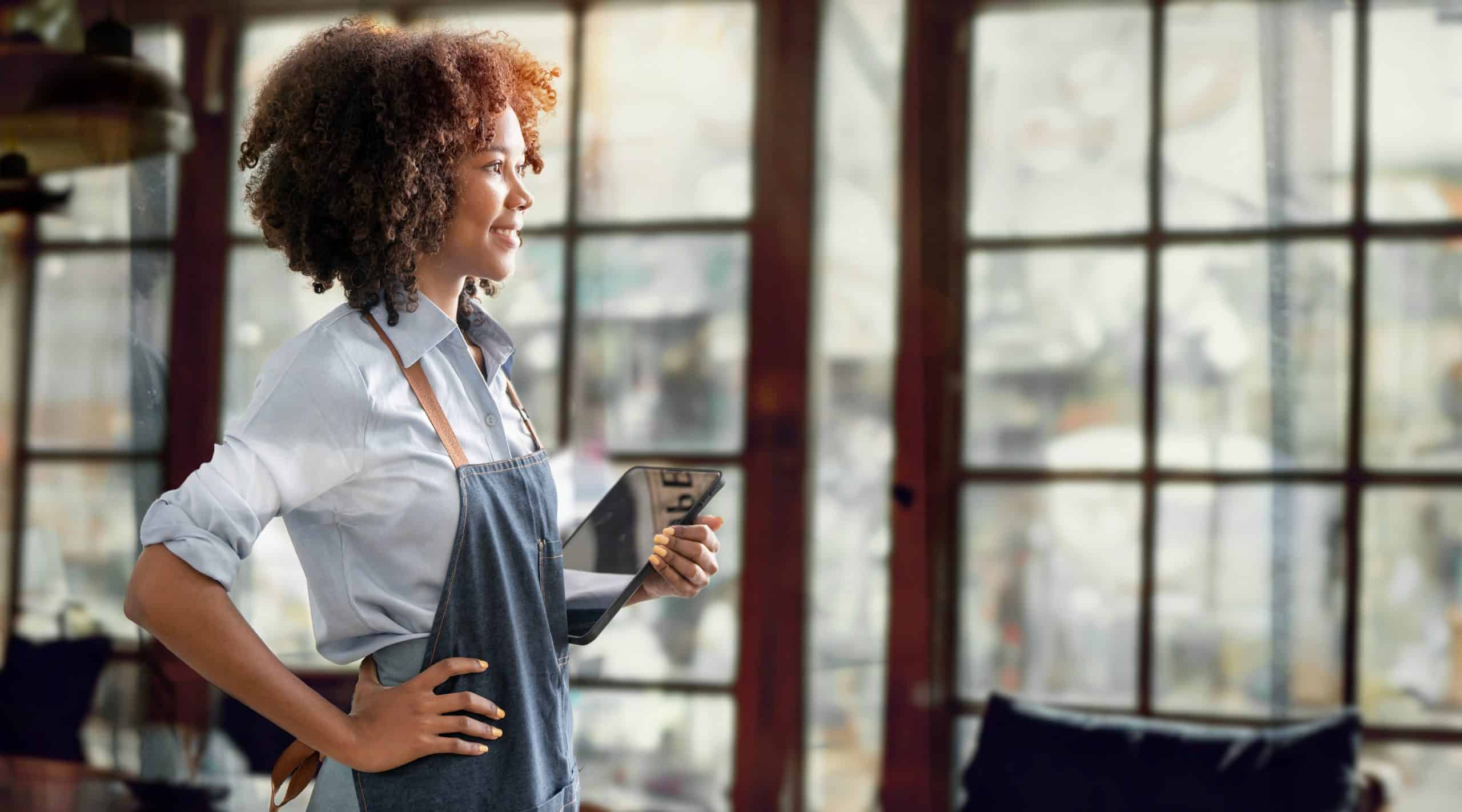 Successful small business owner. Beautiful black skin girl with apron holding tablet standing in coffee shop restaurant. Portrait of latin woman barista cafe owner.