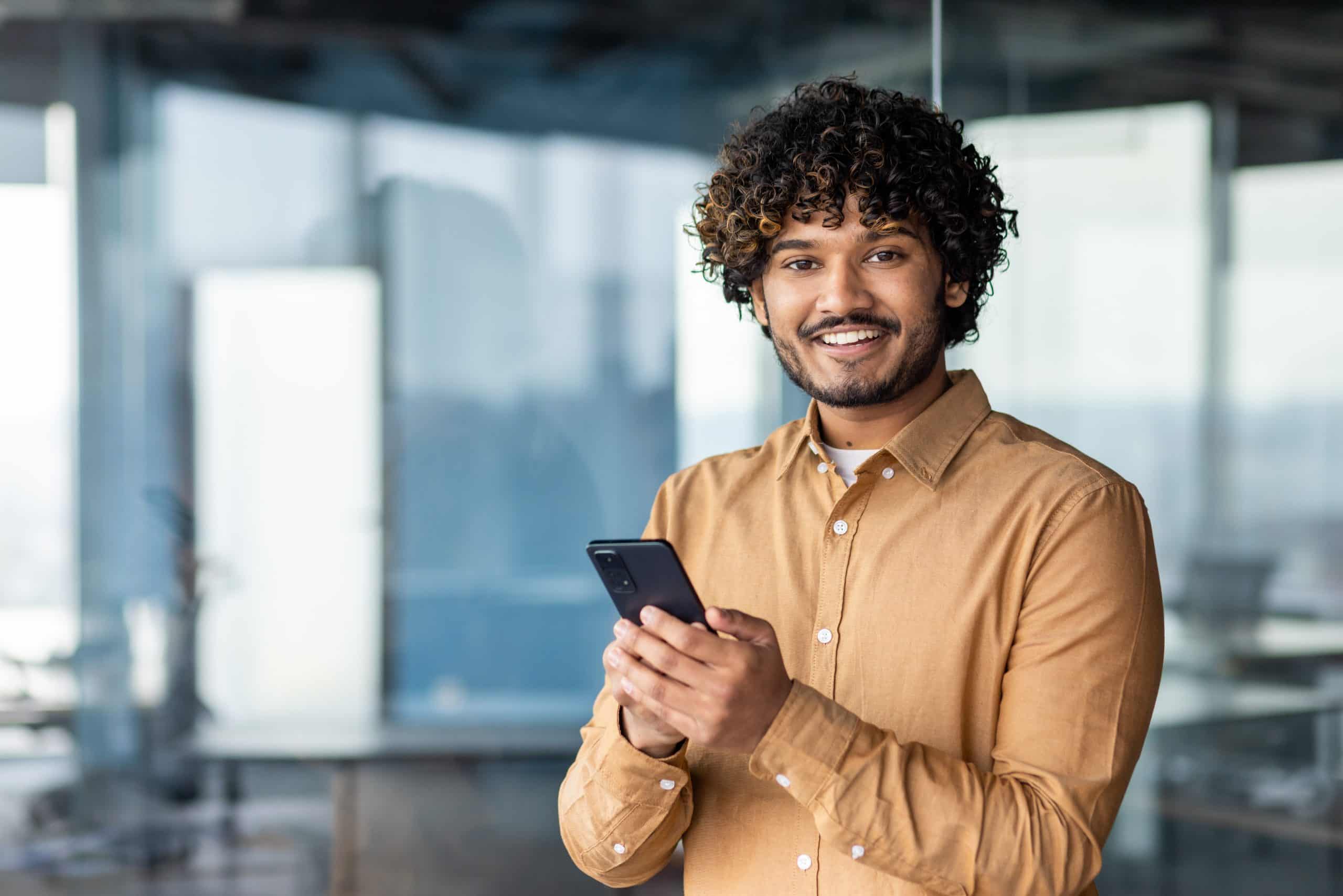 Portrait of a young businessman inside the office, hispanic man is smiling and looking at the camera, the man is holding a phone in his hands, typing a message and browsing the internet
