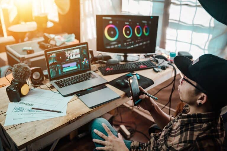 The content creator using mobile working with the laptop editing video footage in the house studio of freelancer man