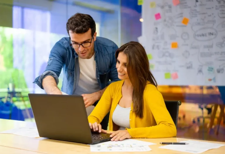 A man and a woman in casual business attire collaborating at a desk with a laptop, in an office with a bright, blurred background and a wall covered in sticky notes and brainstorming diagrams.